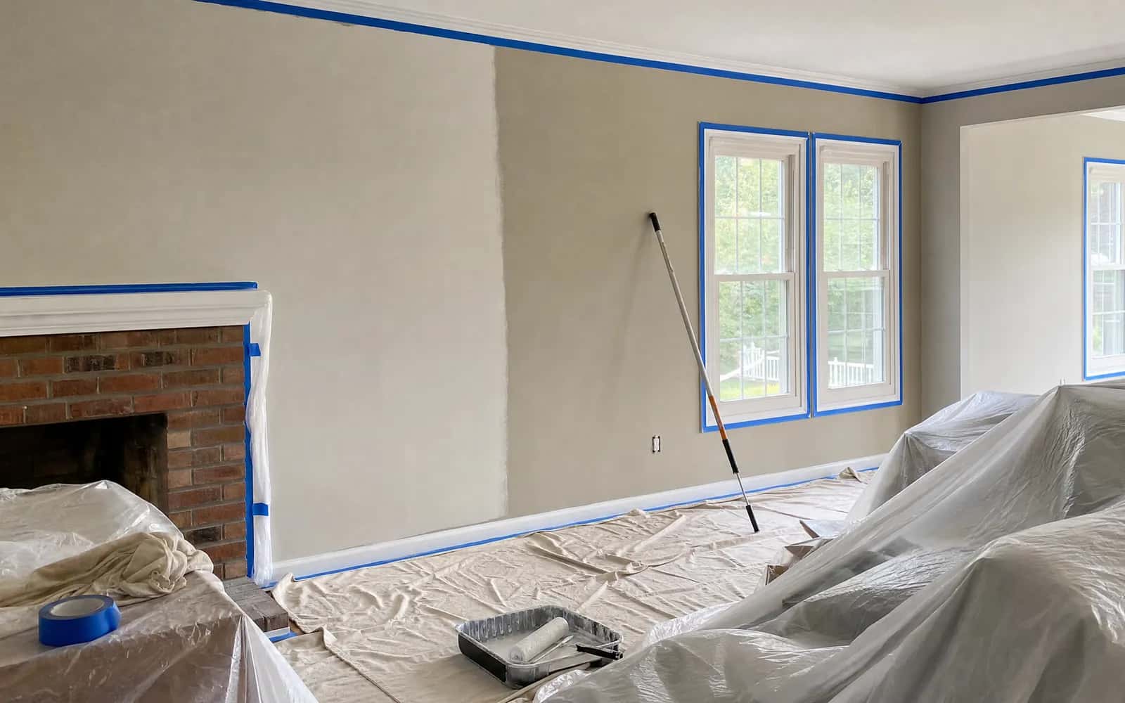 Living room prepped for painting with tape, drop cloths, and a first coat in progress
